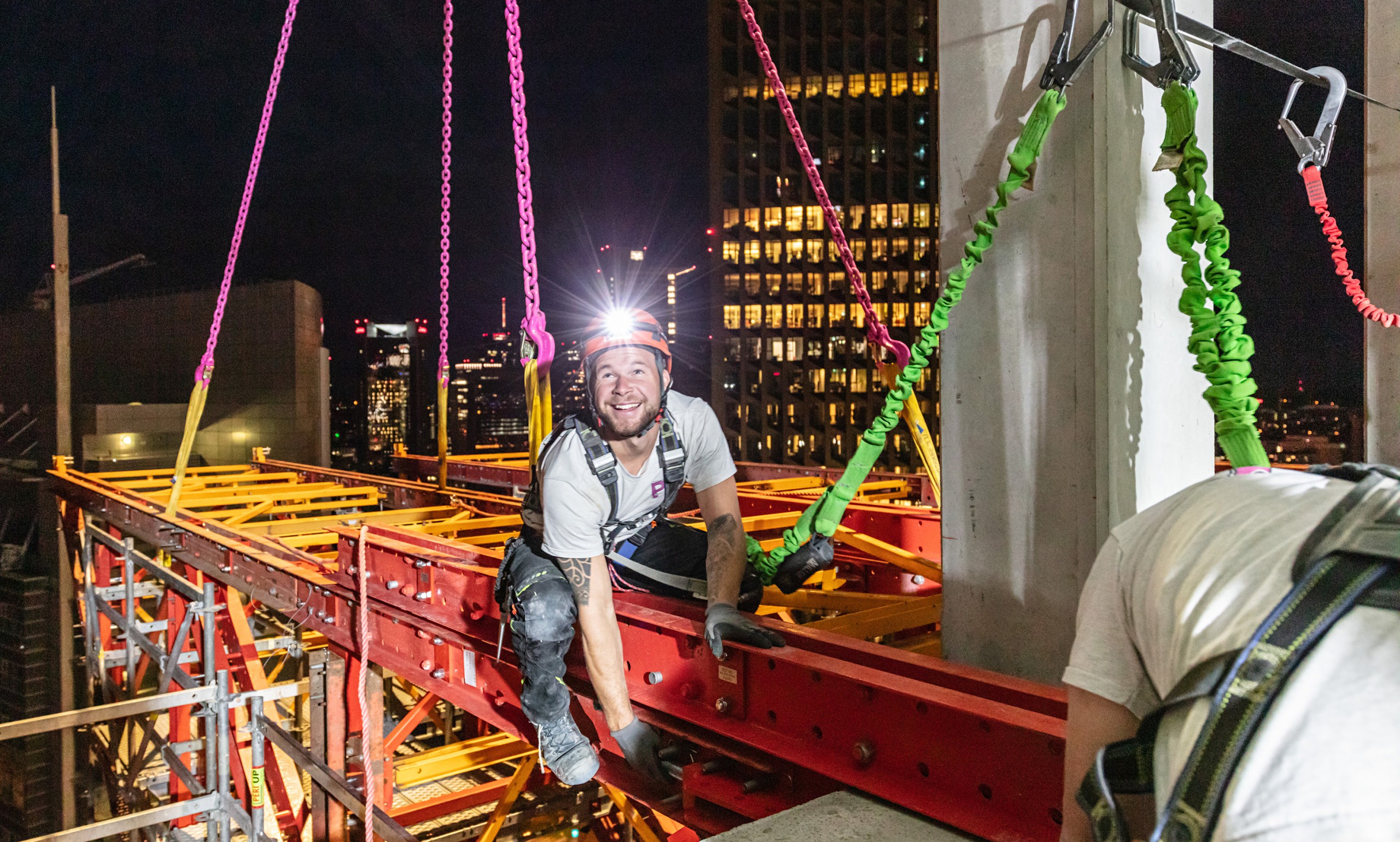 A worker sits on scaffolding at a high-rise construction site. He is secured with straps and is installing a steel beam. The headlamp on his helmet creates a star-shaped glare spot.